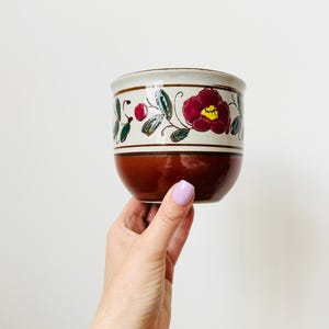 Könnte beinhalten: Eine Keramikschale mit floralem Design. Die Schale hat einen braunen Boden, ein cremefarbenes Band mit floralem Muster und einen cremefarbenen Rand. Das florale Muster umfasst rote Blumen mit gelben Zentren und grüne Blätter.
