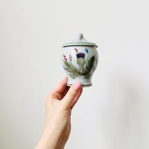 May include: A small, round ceramic jar with a lid, featuring a hand-painted thistle design in shades of purple, green, and blue. The jar has a light blue base and lid, and a small handle on top. A decorative item.