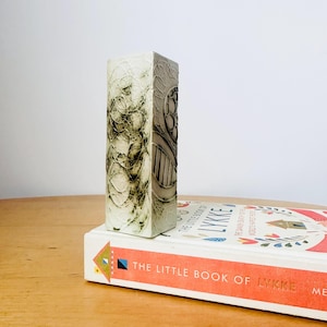 May include: A white ceramic vase with a textured, abstract design in shades of green and grey. The vase is standing on a book titled "The Little Book of Lykke".