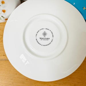 May include: A white fine bone china saucer with a central recessed area. The saucer has the text "WHITTTARD OF CHELSEA" and "MADE IN ENGLAND" printed on the bottom. The saucer is microwave and dishwasher safe.