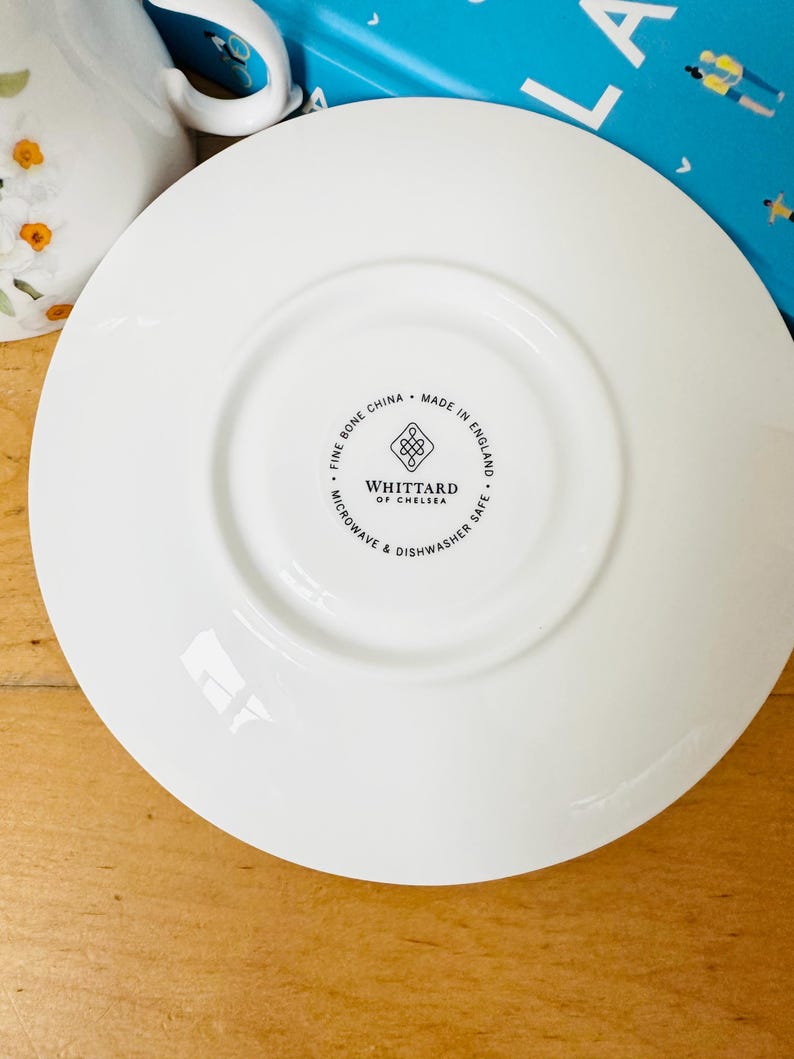 May include: A white fine bone china saucer with a central recessed area. The saucer has the text "WHITTTARD OF CHELSEA" and "MADE IN ENGLAND" printed on the bottom. The saucer is microwave and dishwasher safe.