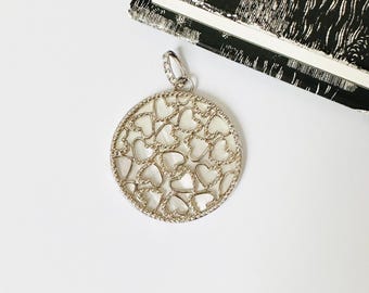 May include: A silver-toned pendant with a heart-shaped design. The pendant has a circular shape with a textured border and a bail adorned with small crystals. The interior features a pattern of interconnected hearts, creating an openwork design.