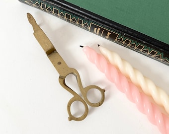 May include: An antique brass candle snuffer with scissor handles rests beside two twisted candles, one pink and one white. A green book with gold accents is in the background. The items are on a white surface.