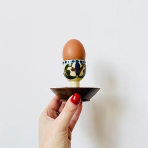 May include: A brown egg sits in a decorative egg cup with a matching saucer. The egg cup is cream-colored with blue, green, and brown floral designs. The saucer is a dark brown color.
