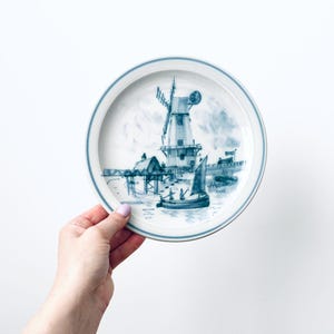 May include: A round, white plate with a blue rim and a detailed blue illustration of a windmill, buildings, and a sailboat on water. The plate is held against a white background.
