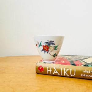 May include: A small white ceramic cup with a floral design in red, yellow, green, and blue. The cup is sitting on top of a book titled "HAIKU" with the text "edited by Jackie  Ha"