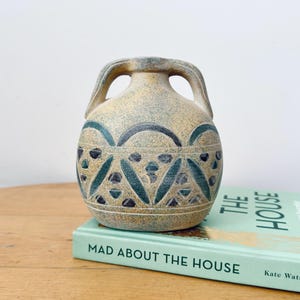 May include: A ceramic vase with two handles, featuring a speckled beige finish and a dark green geometric pattern of arches and triangles around the center. The vase sits atop a light green book titled Mad About The House by Kate Watson-Smyth.