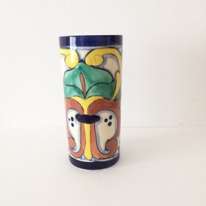 May include: A cylindrical ceramic vase with a blue, yellow, green and red floral design. The vase is hand-painted in a traditional Mexican style.