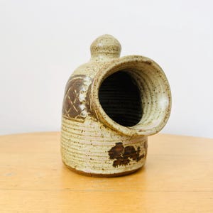 May include: A speckled beige ceramic salt pig with a wide, circular side opening and a small knob handle on top. The exterior features brown, hand-painted geometric cross-hatch patterns and dark brown splotches over a textured, light-colored glaze.