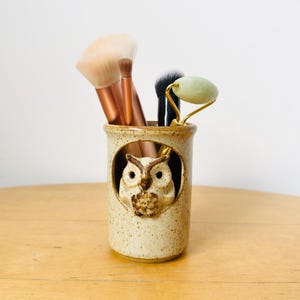 May include: A beige ceramic owl-themed holder with makeup brushes and a jade roller. The holder has a detailed owl design and is filled with various makeup brushes and a jade roller. The holder is on a wooden surface.
