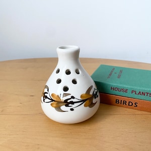 May include: White ceramic vase with a floral design in brown and gold. The vase has holes around the middle for holding flowers.