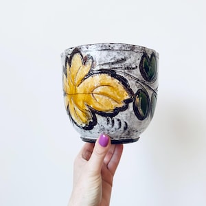 May include: A ceramic bowl with a white, grey, and black speckled exterior. The bowl features a large yellow leaf design with black outlines and green leaf accents. The bowl is held by a hand.