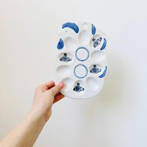 Puede incluir: Un porta huevos de cerámica blanca con forma de gallina con diseños florales azules y blancos. El porta huevos tiene 12 compartimentos individuales con forma de huevo.