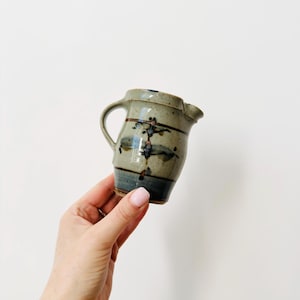 Puede incluir: Un pequeño jarro de cerámica hecho a mano, con asa y boquilla. El jarro presenta una base beige moteada con diseños abstractos azules y marrones. La parte inferior es azul oscuro. Una pieza única para servir.