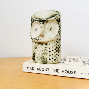 Może przedstawiać: Ceramiczna figurka sowy w kremowym kolorze z czarnymi detalami. Sowa ma duże, okrągłe oczy i stoi na książce zatytułowanej "MAD ABOUT THE HOUSE". Figurka jest przedmiotem dekoracyjnym.