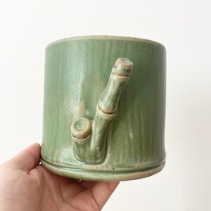 May include: A green ceramic mug with a bamboo-shaped handle. The mug has a textured, slightly speckled surface.