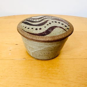 Può includere: Un vaso in ceramica fatto a mano con coperchio. Il vaso è di colore beige chiaro con un bordo marrone e un coperchio decorato con un motivo a onde marroni. Il vaso è appoggiato su una superficie di legno.