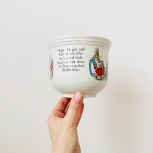 Puede incluir: Cuenco de cerámica blanca con ilustraciones de conejos y texto de un cuento infantil. El cuenco tiene forma redondeada con un borde ligeramente ensanchado. El texto dice: "Flops, Mopsy, and Cottontail, who were good little bunnies, went down the lane to gather blackberries:"