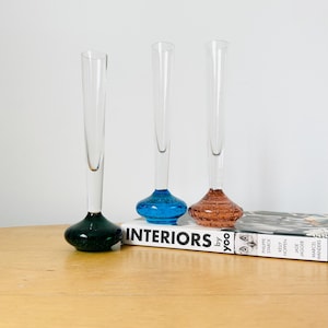 May include: Three elegant glass vases with coloured bases: dark green, blue, and brown. The vases are clear with long, narrow necks. The bases have a rounded shape. The vases are displayed on a wooden surface next to a book titled "INTERIORS".