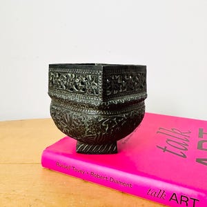 May include: An antique, dark metal container with detailed floral patterns. The container features a square top and a rounded base, supported by a small, square foot. It is placed on a vibrant pink book with the title "talk ART".