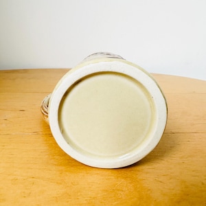 Op de afbeelding: De onderkant van een keramische mok met een lichtbruine glazuur. De mok heeft een ronde bodem en een licht verhoogde lip.