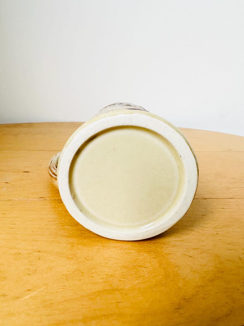 Op de afbeelding: De onderkant van een keramische mok met een lichtbruine glazuur. De mok heeft een ronde bodem en een licht verhoogde lip.