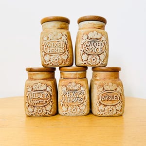 Puede incluir: Seis botes de especias vintage con tapas de madera. Los botes beige presentan diseños florales en relieve y etiquetas que dicen "Mixed Herbs", "Mixed Spice", "Black Peppercorns", "Bay Leaves" y "Parsley". Están colocados sobre una superficie marrón claro.