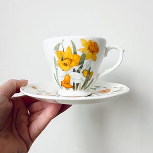 May include: White teacup and saucer set featuring a floral design of yellow daffodils and green leaves. The teacup has a curved handle and the saucer is round. The set is likely made of porcelain or ceramic.