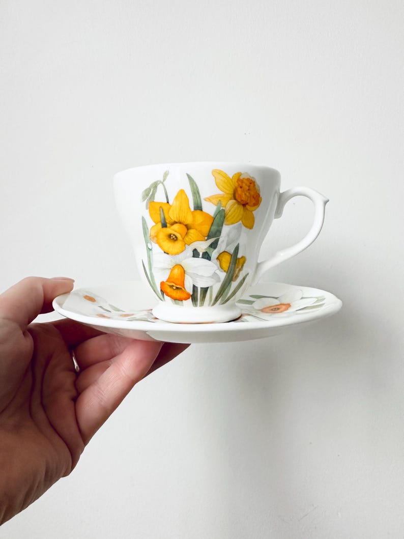 May include: White teacup and saucer set featuring a floral design of yellow daffodils and green leaves. The teacup has a curved handle and the saucer is round. The set is likely made of porcelain or ceramic.