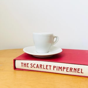 May include: A white porcelain teacup and saucer sitting on top of a red book with the title "The Scarlet Pimpernel" printed in white.