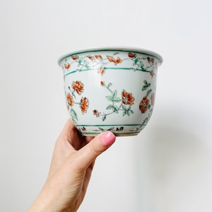 May include: A white ceramic planter with a floral design. The planter features orange flowers and green leaves, with a green border around the rim. The planter is held by a hand.