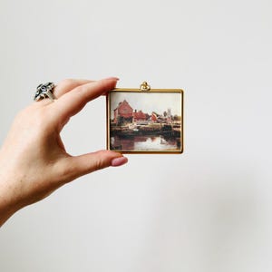 May include: A small, square, gold-toned picture frame holding a landscape painting of buildings along a river. The painting features red-roofed buildings, a church, and a body of water. The frame is held by a hand against a white background.