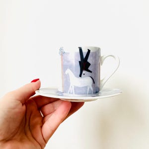 May include: A white ceramic teacup and saucer set. The cup features a whimsical design of a white horse and a figure in black against a blue and purple background. The saucer has a matching blue and white pattern.