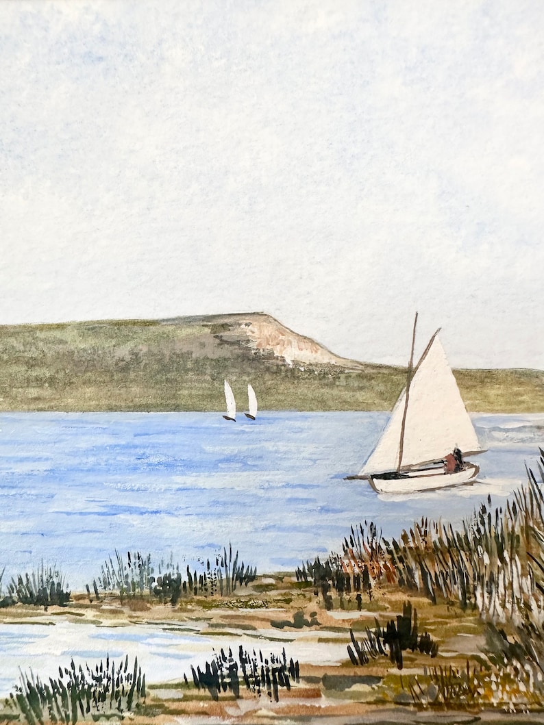 Puede incluir: Una pintura de acuarela de un velero en una masa de agua con una colina verde en el fondo. El velero tiene una vela blanca y navega hacia la derecha de la imagen. Hay dos veleros m&aacute;s peque&ntilde;os en la distancia.