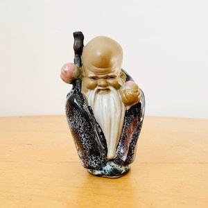 Könnte beinhalten: Keramikfigur eines weisen, alten Mannes mit langem, weißem Bart, der einen Stab und einen Pfirsich hält. Die Figur trägt eine schwarze Robe mit gesprenkeltem Muster und hat einen ruhigen Ausdruck. Dekorativer Artikel.