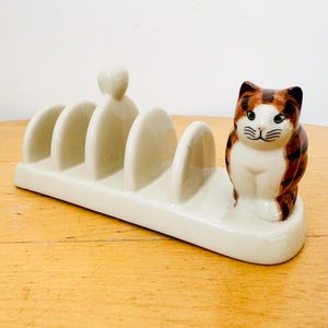 May include: Cream-colored ceramic toast rack with a decorative cat figurine. The rack has five slots for toast and a small cat figurine with brown and white markings. The cat has green eyes and is sitting upright.