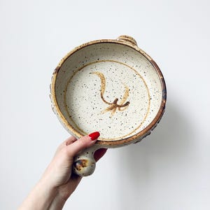 May include: A speckled ceramic serving dish with a handle. The dish has a light beige interior with a brown floral design and a dark brown rim. The handle is a rounded shape. The dish is held against a white background.