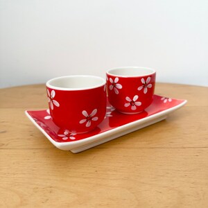 May include: Two red ceramic egg cups with white floral designs, sitting on a white rectangular tray. The tray is on a wooden surface.
