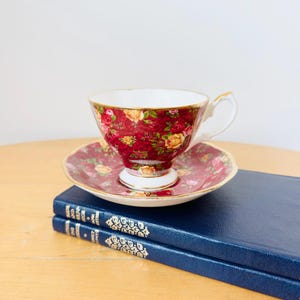 Puede incluir: Un juego de taza y platillo vintage con un estampado floral en rojo, amarillo y rosa. La taza tiene un asa blanca y ribetes dorados. El juego descansa sobre dos libros azules.