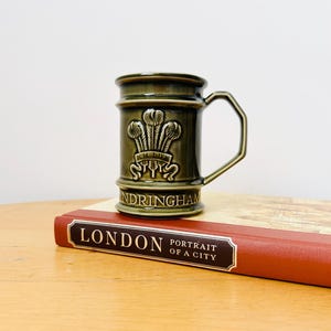 May include: A green ceramic mug with a white crest and the words "Ich Dien" and "Andringham" on the side. The mug is sitting on top of a red book with the title "London Portrait of a City".