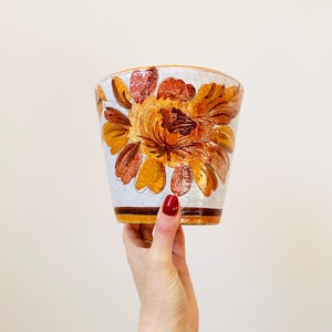 Op de afbeelding: Een witte keramische plantenpot met een bloemenontwerp in tinten oranje, bruin en geel. De pot heeft een brede rand en een licht uitlopende basis.