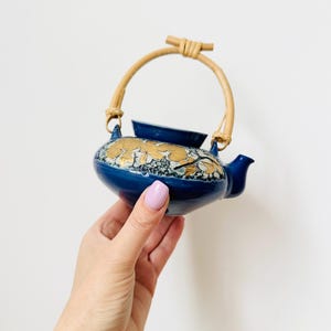 May include: A small, navy blue ceramic teapot with a bamboo handle. The teapot features a gold floral design around the middle. The spout and lid are also navy blue. The teapot is held in a person's hand.
