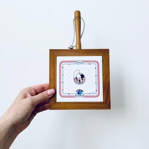 May include: A square wooden trivet with a ceramic tile featuring a cartoon character in a chef's hat. The tile has a red and blue border with a polka dot background. A wooden handle is attached.