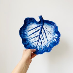 May include: A blue and white ceramic serving dish shaped like a cabbage leaf. The dish has a detailed design of veins and a white centre.