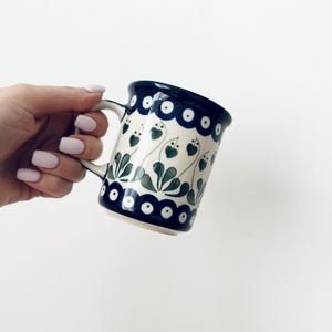 Pode incluir: Uma caneca de cerâmica com base na cor creme, apresentando uma borda superior e inferior azul-escura decorada com pontos brancos. O corpo da caneca exibe um padrão repetitivo de formas de folhas verdes e caules finos.