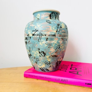 Può includere: Un vaso in ceramica con base azzurra, decorato con disegni astratti neri, rosa e crema. Il vaso ha un corpo arrotondato e un bordo leggermente svasato. È posto su una superficie di legno accanto a un libro rosa intitolato "talk ART".