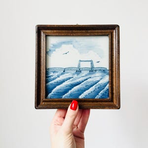 May include: A framed artwork featuring a blue and white depiction of a bridge over water, with birds in the sky. The artwork is in a square, dark wood frame.