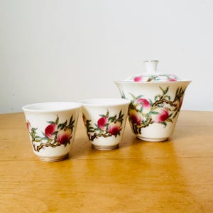 Puede incluir: Un juego de té de porcelana blanca con un Gaiwan con tapa y dos tazas pequeñas. Cada pieza está decorada con una rama de melocotones en tonos rojos, rosas y verdes. El juego se exhibe sobre una superficie de madera.