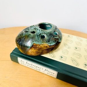 May include: A green and brown ceramic bud vase with a rounded shape and multiple holes for flowers. The vase is sitting on top of a green book titled "An Innkeeper's Diary" by John  Fothergill  Fatio.