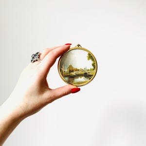 May include: A round, gold-toned frame holds a miniature landscape painting. The painting depicts a waterside scene with buildings, trees, and a bridge. The frame is held by a hand with red nail polish and a silver ring.